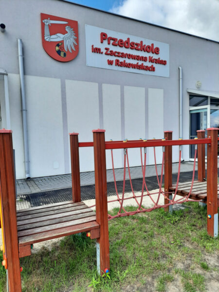 Ściana boczna budynku przedszkola z widocznym czerwonym napisem "Przedszkole im. Zaczarowana Kraina w Rakowiskach". Na pierwszym planie plac zabaw.