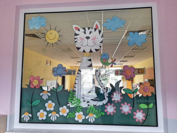 Przeszklenie między korytarze a jedną z sal. Na przeszkleniu naklejone ozdoby przedstawiające kota, słońce, chmury i kwiaty