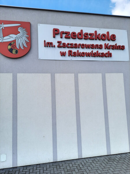 Ściana boczna budynku przedszkola z widocznym czerwonym napisem "Przedszkole im. Zaczarowana Kraina w Rakowiskach".